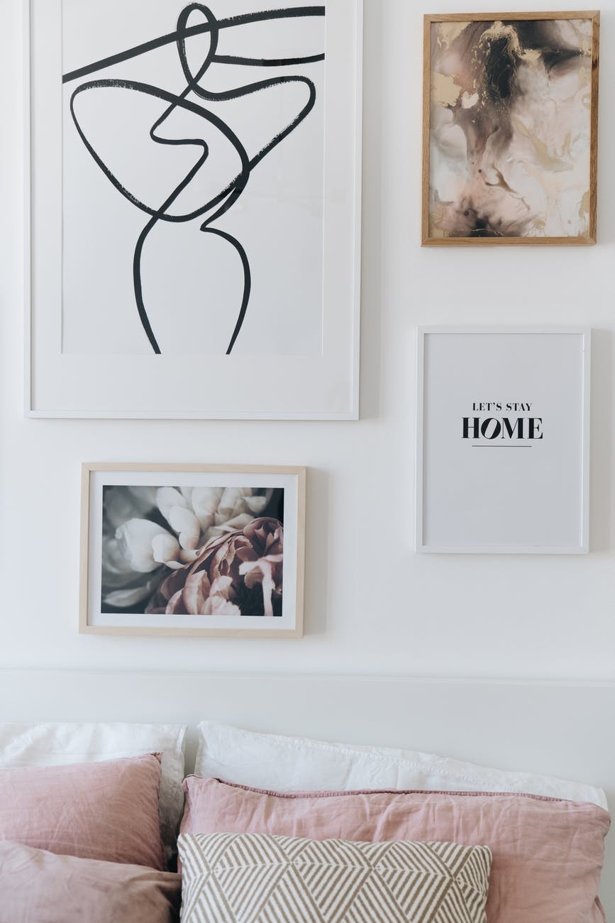 white wall with white and pink pillows on bed, white framed art with black abstract art and art with flower photography on wall , Let's stay home Black letters on white wall art in white frame